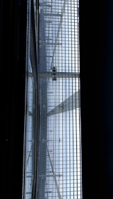 A view of a metal gratin with a person walking on it, seen from below. The structure consists of a grid of metal bars, allowing light to pass through and casting shadows onto the surface below. The scene has a geometric, industrial feel.