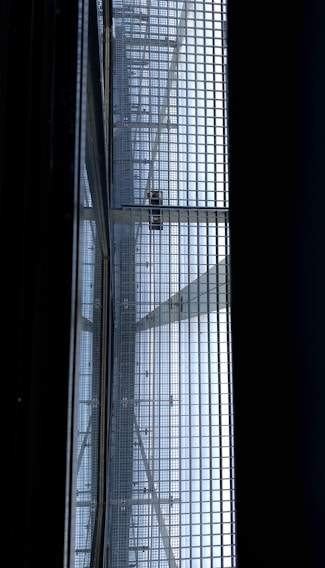 A view of a metal gratin with a person walking on it, seen from below. The structure consists of a grid of metal bars, allowing light to pass through and casting shadows onto the surface below. The scene has a geometric, industrial feel.