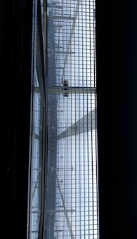 A view of a metal gratin with a person walking on it, seen from below. The structure consists of a grid of metal bars, allowing light to pass through and casting shadows onto the surface below. The scene has a geometric, industrial feel.