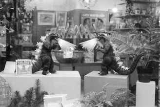 Two toy figures resembling dinosaurs or monsters are positioned facing each other on display pedestals. They have spiky backs and are emitting light beams from their mouths. The scene is set within a store window decorated with ferns and other plants in pots.