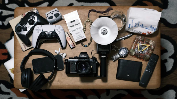 A tabletop surface is filled with various items, including two gaming controllers, a pack of cigarettes, a smartphone, headphones, a camera, a watch, a glass with liquid, a remote control, a notepad, and an ashtray with cigarettes. These are neatly arranged on the wooden table.