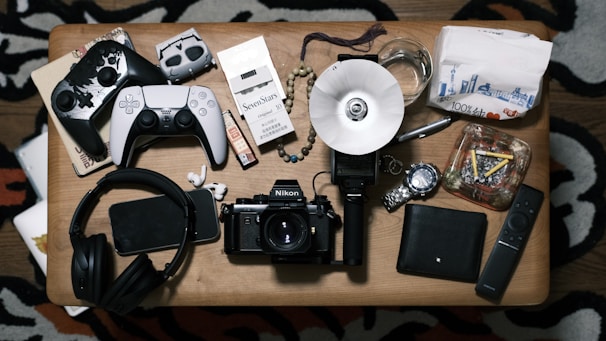 A tabletop surface is filled with various items, including two gaming controllers, a pack of cigarettes, a smartphone, headphones, a camera, a watch, a glass with liquid, a remote control, a notepad, and an ashtray with cigarettes. These are neatly arranged on the wooden table.