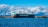 A large cargo ship with the name 'WALLENIUS WILHELMSEN' is prominently positioned on calm blue waters. The background features a scenic view with expansive clouds and distant mountains under a clear sky. The shoreline is lined with trees, and there is a tall communication tower visible to the left.