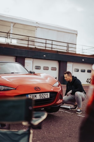 A man crouches beside a red Mazda car, which is parked in front of a garage with multiple doors. He seems focused, possibly working on or inspecting the car. A chair is in the foreground, slightly out of focus.