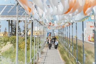 A group of people walks along a pathway decorated with orange and white fabric banners overhead. The path is lined with greenery on one side and grass on the other. Solar panels are visible on a nearby structure, and there are signs with text in the background.
