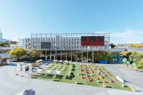 A large open outdoor area with seating arrangements consisting of colorful bean bags spread out on a green artificial grass surface. Several shaded structures with white canopies are set up, and a gray multi-story building with multiple windows serves as a backdrop. The building features two large digital billboards, one dark and one red. Surrounding the area are trees and some small green structures that appear to be decorative elements or seating. The sky is clear and bright.