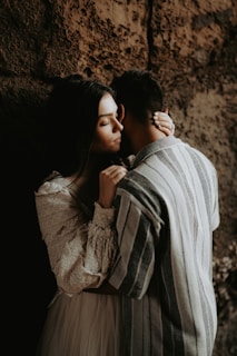 A couple stands closely together in an intimate embrace, with one person resting their head on the other's shoulder. They are positioned against a textured, earthy background. The lighting is soft and muted, casting gentle shadows.