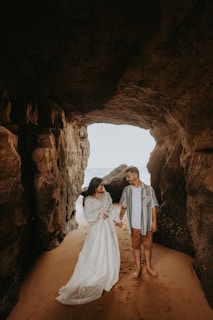 A couple stands inside a rocky cave opening that leads to the sea. The woman is wearing a long white dress, and the man is dressed in a casual striped shirt and shorts. They are holding hands, surrounded by the warm tones of the cave walls and the sandy floor.