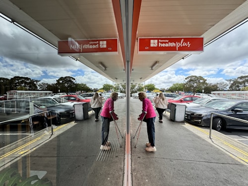 A reflection scene outside a healthcare facility with a red sign reading 'Ramsay health plus'. An elderly person is walking with crutches, wearing a pink jacket. Multiple parked cars are visible along with some trees in the background. The sky is partially cloudy.