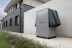A modern, rectangular air conditioning or heating unit with the logo 'alpha innotec' is installed outside a building with a dark gray brick exterior. The unit is positioned against a light concrete wall, and there is overgrown grass along the edge of the pavement.