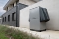A modern, rectangular air conditioning or heating unit with the logo 'alpha innotec' is installed outside a building with a dark gray brick exterior. The unit is positioned against a light concrete wall, and there is overgrown grass along the edge of the pavement.