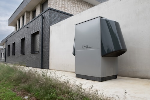 A modern, rectangular air conditioning or heating unit with the logo 'alpha innotec' is installed outside a building with a dark gray brick exterior. The unit is positioned against a light concrete wall, and there is overgrown grass along the edge of the pavement.