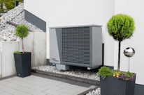 An outdoor area features a modern heat pump unit positioned against a white wall. The setting is minimalist, with a stone and pebble landscape surrounding the unit. Two black planters contain manicured green shrubs, adding a touch of nature to the space. A reflective silver garden sphere provides a decorative element amidst the functional design.