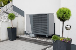 An outdoor area features a modern heat pump unit positioned against a white wall. The setting is minimalist, with a stone and pebble landscape surrounding the unit. Two black planters contain manicured green shrubs, adding a touch of nature to the space. A reflective silver garden sphere provides a decorative element amidst the functional design.