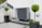 An outdoor area features a modern heat pump unit positioned against a white wall. The setting is minimalist, with a stone and pebble landscape surrounding the unit. Two black planters contain manicured green shrubs, adding a touch of nature to the space. A reflective silver garden sphere provides a decorative element amidst the functional design.