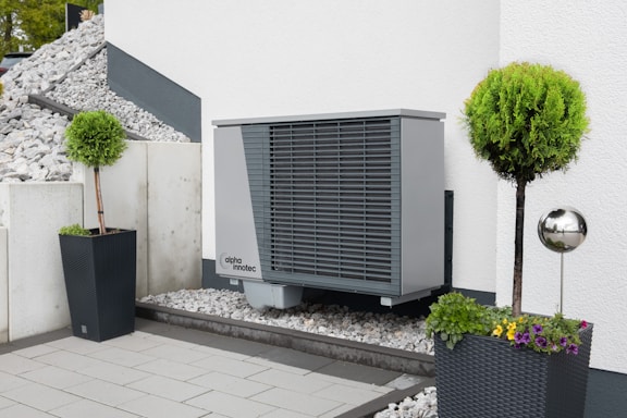 An outdoor area features a modern heat pump unit positioned against a white wall. The setting is minimalist, with a stone and pebble landscape surrounding the unit. Two black planters contain manicured green shrubs, adding a touch of nature to the space. A reflective silver garden sphere provides a decorative element amidst the functional design.