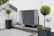 An outdoor area features a modern heat pump unit positioned against a white wall. The setting is minimalist, with a stone and pebble landscape surrounding the unit. Two black planters contain manicured green shrubs, adding a touch of nature to the space. A reflective silver garden sphere provides a decorative element amidst the functional design.