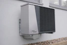 A modern, wall-mounted exterior heat pump unit with a sleek, metallic design is securely attached to the side of a building. The unit features a prominent black grille on its front and is situated above a layer of small rocks bordering the building. This setting suggests installation for heating or cooling purposes.