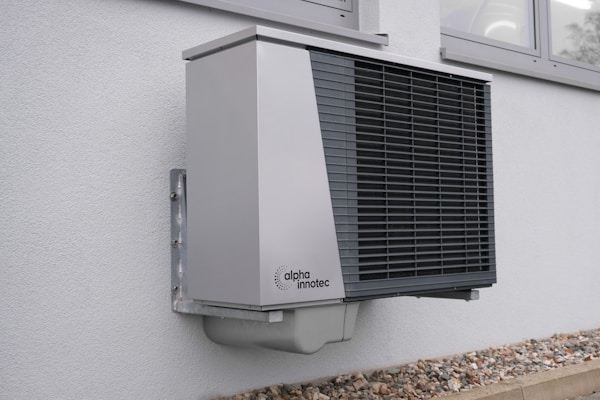 A modern, wall-mounted exterior heat pump unit with a sleek, metallic design is securely attached to the side of a building. The unit features a prominent black grille on its front and is situated above a layer of small rocks bordering the building. This setting suggests installation for heating or cooling purposes.
