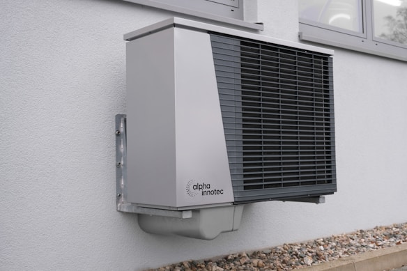 A modern, wall-mounted exterior heat pump unit with a sleek, metallic design is securely attached to the side of a building. The unit features a prominent black grille on its front and is situated above a layer of small rocks bordering the building. This setting suggests installation for heating or cooling purposes.