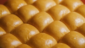 A close-up view of several rows of freshly baked golden brown bread rolls, each with a smooth, shiny surface that suggests a soft texture.