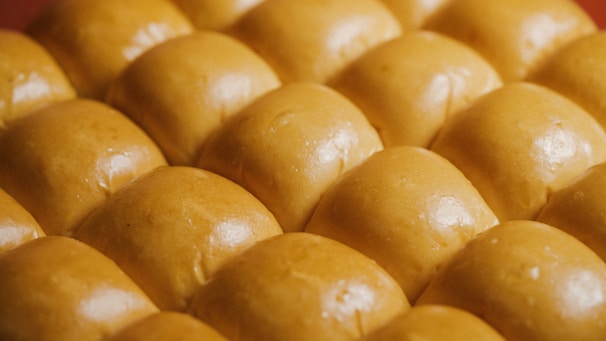 A close-up view of several rows of freshly baked golden brown bread rolls, each with a smooth, shiny surface that suggests a soft texture.