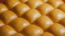 A close-up view of several rows of freshly baked golden brown bread rolls, each with a smooth, shiny surface that suggests a soft texture.