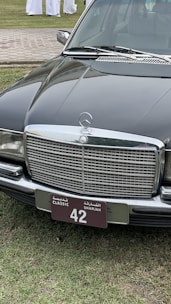 A classic black Mercedes-Benz car is parked on a grassy area. The focus is on the car's front, showcasing a distinctive chrome grille and a license plate with 'Classic Sharjah 42'. The background includes a paved path and several people wearing white garments.