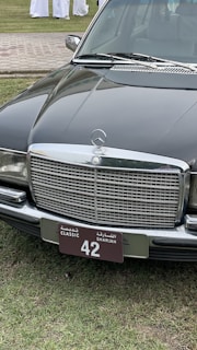 A classic black Mercedes-Benz car is parked on a grassy area. The focus is on the car's front, showcasing a distinctive chrome grille and a license plate with 'Classic Sharjah 42'. The background includes a paved path and several people wearing white garments.