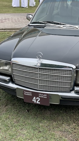 A classic black Mercedes-Benz car is parked on a grassy area. The focus is on the car's front, showcasing a distinctive chrome grille and a license plate with 'Classic Sharjah 42'. The background includes a paved path and several people wearing white garments.