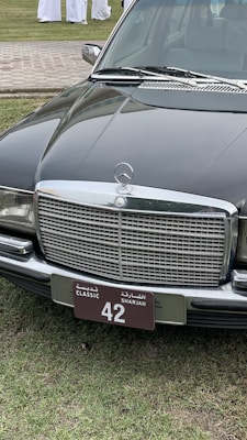 A classic black Mercedes-Benz car is parked on a grassy area. The focus is on the car's front, showcasing a distinctive chrome grille and a license plate with 'Classic Sharjah 42'. The background includes a paved path and several people wearing white garments.