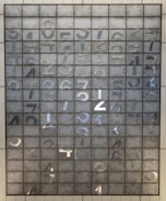 An array of metal plates arranged in a grid pattern, each square featuring a metallic numeral from 0 to 9. The numbers are reflective and appear to be stamped or cut out. The background shows a matte gray texture, adding contrast to the shiny metallic characters.