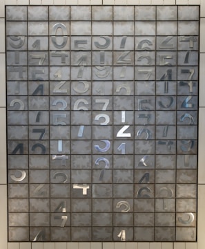 An array of metal plates arranged in a grid pattern, each square featuring a metallic numeral from 0 to 9. The numbers are reflective and appear to be stamped or cut out. The background shows a matte gray texture, adding contrast to the shiny metallic characters.