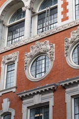 A building facade adorned with intricate architectural details. The structure features large arched windows with decorative stonework, including floral and facial motifs. A ladder is positioned against one of the upper windows, adding a modern element to the classic design. The main material appears to be red brick with white stone accents, providing a striking contrast.