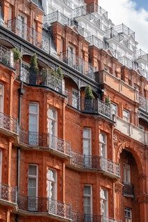 A grand architectural facade featuring ornate wrought iron balconies and deep red brickwork. The building showcases multiple stories with numerous windows, adorned with intricate detailing. Some greenery is visible on the balconies, adding a touch of nature to the urban setting.