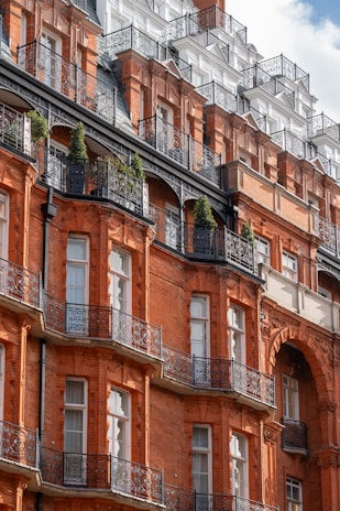 A grand architectural facade featuring ornate wrought iron balconies and deep red brickwork. The building showcases multiple stories with numerous windows, adorned with intricate detailing. Some greenery is visible on the balconies, adding a touch of nature to the urban setting.