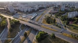Aerial view of a cityscape featuring multiple intersecting highways surrounded by green spaces and residential buildings. Several cars are traveling on the roads, and apartment complexes are visible in the background. The area appears organized with defined lanes and pathways, and the urban environment is interspersed with trees and grassy areas.