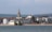 A coastal townscape features a prominent church spire surrounded by rows of traditional buildings. In the foreground, there is a sandy beach next to a large body of water. Rolling green hills extend into the background, suggesting a serene and picturesque setting.