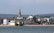 A coastal townscape features a prominent church spire surrounded by rows of traditional buildings. In the foreground, there is a sandy beach next to a large body of water. Rolling green hills extend into the background, suggesting a serene and picturesque setting.