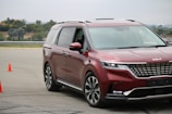 A shiny red minivan navigates a driving course marked by two bright orange cones on an asphalt surface. The vehicle displays modern styling with sleek headlights and a prominent front grille. Trees and a fence are visible in the background under a cloudy sky.