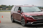A shiny red minivan navigates a driving course marked by two bright orange cones on an asphalt surface. The vehicle displays modern styling with sleek headlights and a prominent front grille. Trees and a fence are visible in the background under a cloudy sky.