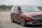 A shiny red minivan navigates a driving course marked by two bright orange cones on an asphalt surface. The vehicle displays modern styling with sleek headlights and a prominent front grille. Trees and a fence are visible in the background under a cloudy sky.