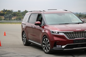 A shiny red minivan navigates a driving course marked by two bright orange cones on an asphalt surface. The vehicle displays modern styling with sleek headlights and a prominent front grille. Trees and a fence are visible in the background under a cloudy sky.