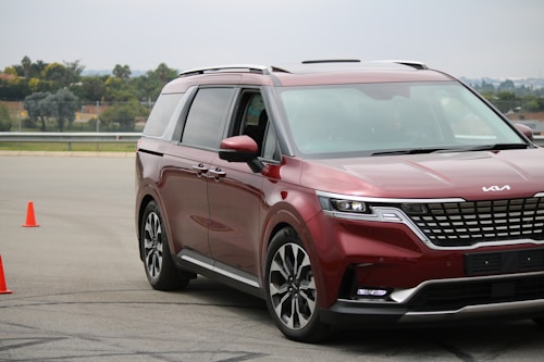 A shiny red minivan navigates a driving course marked by two bright orange cones on an asphalt surface. The vehicle displays modern styling with sleek headlights and a prominent front grille. Trees and a fence are visible in the background under a cloudy sky.