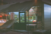 A small kiosk or shop with a corrugated metal roof, seen at night. The interior is dimly lit, with some illuminated signage in the window. The shop's exterior includes a ramp or platform leading up to the entrance. There is signage on the front window with text and images, likely advertising products. Snow can be seen on the ground outside.