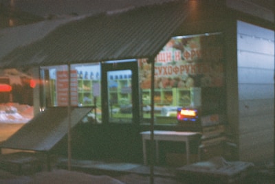 A small kiosk or shop with a corrugated metal roof, seen at night. The interior is dimly lit, with some illuminated signage in the window. The shop's exterior includes a ramp or platform leading up to the entrance. There is signage on the front window with text and images, likely advertising products. Snow can be seen on the ground outside.
