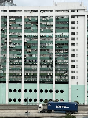 A large building facade is covered with numerous windows, each equipped with air conditioning units. The building features a pattern of alternating green and white sections, and a distinctive row of circular openings at the lower level. In the foreground, a truck with a blue container labeled 'KMTC Line' is parked on a road, and a person on a green motorcycle is nearby.