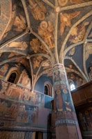 The interior of an ornately decorated church is depicted, featuring a vaulted ceiling and walls adorned with religious frescoes. The art showcases various biblical figures and scenes in muted earth tones, primarily utilizing blues, browns, and golds. The architectural details include pointed arches and intricately painted columns that emphasize the grandeur and historical significance of the space.