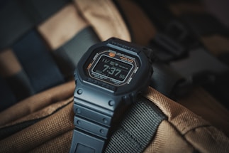 A digital watch with a rugged design, featuring a black strap and a digital display showing the time 7:37. The background consists of a textured, tan and green fabric, possibly part of a backpack or outdoor gear.