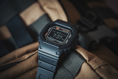 A digital watch with a rugged design, featuring a black strap and a digital display showing the time 7:37. The background consists of a textured, tan and green fabric, possibly part of a backpack or outdoor gear.